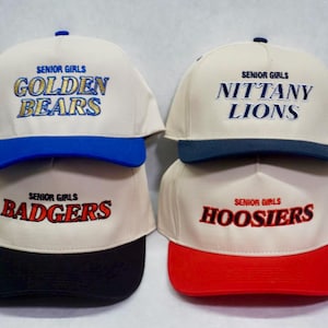 May include: Four white baseball caps with different colored brims and embroidered text. The caps say "Senior Girls Golden Bears", "Senior Girls Nittany Lions", "Senior Girls Badgers", and "Senior Girls Hoosiers".