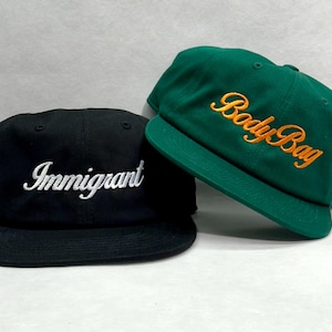 Peut inclure: Deux casquettes de baseball. L'une est noire avec le mot "Immigrant" brodé en blanc. L'autre est verte avec "Body Bag" brodé en orange. Les deux casquettes ont une visière plate et une couronne structurée.