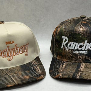 Puede incluir: Dos gorras de béisbol. Una es beige con "BOLA bodybag" bordado en naranja. La otra es camuflaje con "Ranchero Outdoors" en blanco. Ambas tienen una visera de camuflaje y un botón en la parte superior.