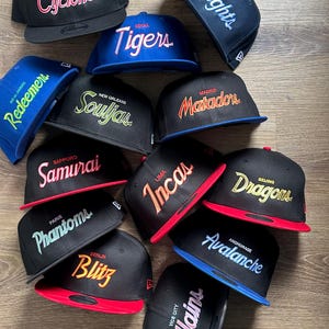 Puede incluir: Una colección de gorras de béisbol en varios colores, con nombres y logotipos de equipos bordados. Las gorras incluyen los nombres "Cyclones", "Tigers", "Knights", "Souljas", "Matadors", "Samurai", "Incas", "Dragons", "Phantoms", "Blitz", "Avalanche" y "Villains".