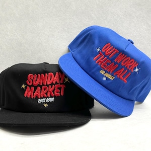 May include: Two baseball caps. One black cap with red embroidered text reading "SUNDAY MARKET ROSE BOWL". The other is a blue cap with red embroidered text that says "OUT WORK THEM ALL LOS ANGELES".