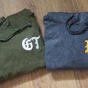 May include: Two folded hoodies, one olive green with white "GT" lettering, and one dark blue with yellow "FP" lettering. Both hoodies have drawstrings and are made of a soft, textured fabric. The hoodies are displayed on a wooden surface.