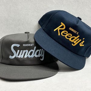 May include: Two baseball caps. One is grey with "MARKET Sunday" embroidered in light blue. The other is navy blue with "ROBERT E. Reedy's" embroidered in yellow. Both caps have a flat peak and adjustable strap.