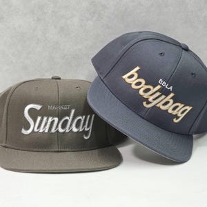 May include: Two baseball caps, one olive green with white embroidered text reading "Sunday MARKET" and one dark grey with gold embroidered text reading "bodybag BBLA". Both caps feature a structured crown, a flat brim, and eyelet vents.