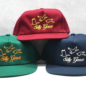 May include: Three baseball caps in different colors: red, green, and blue. Each cap features a white embroidered design of geese flying with the text "Silly Goose" underneath.