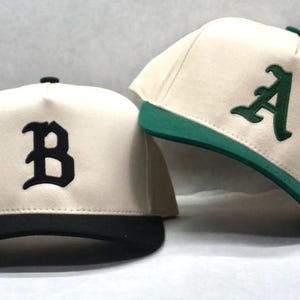 May include: Two baseball caps with a white crown and a contrasting brim. One cap has a black brim and a black letter "B" on the front. The other cap has a green brim and a green letter "A" on the front.