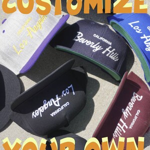 Puede incluir: Una colección de seis gorras de béisbol negras, grises, azules y granate con texto bordado en blanco que dice "CALIFORNIA" y "Los Ángeles" o "Beverly Hills". Las gorras están dispuestas sobre una superficie marrón claro. El texto "CUSTOMIZE YOUR OWN" está escrito en letras naranjas en la parte superior de la imagen.