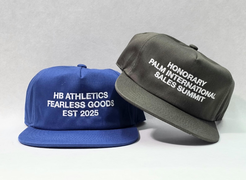 May include: Two flat-brimmed caps. One is navy blue with white text reading "HB ATHLETICS FEARLESS GOODS EST 2025." The other is gray with white text that says "HONORARY PALM INTERNATIONAL SALES SUMMIT."