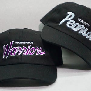 Puede incluir: Dos gorras de béisbol negras con logotipos bordados. La primera gorra tiene "WARRENTON WARRIORS" en letras moradas y blancas. La segunda gorra tiene "TORONTO PEARSON" en letras blancas.