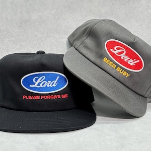 Może przedstawiać: Dwie czapki bejsbolowe. Jedna czarna czapka z napisem "Lord" w niebieskim owalu z tekstem "PLEASE FORGIVE ME" poniżej. Druga jest szara z czerwonym owalem z napisem "Devil" i "BEEN BUSY".