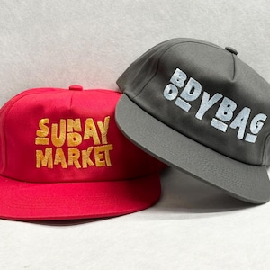 May include: Two flat-brimmed caps. One is red with "SUNDAY MARKET" in gold lettering. The other is grey with "BODYBAG" in white lettering. Both caps have a structured, six-panel design with a flat brim.