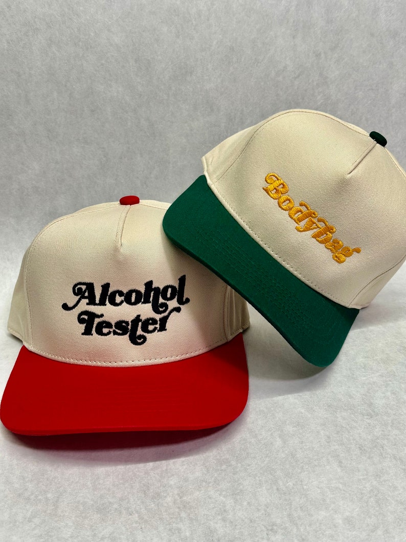 May include: Two baseball caps with different coloured brims. The cap on the left has a red brim and says "Alcohol Tester" in black script. The cap on the right has a green brim and says "Boozy" in gold script.