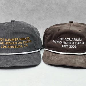 May include: Two corduroy baseball caps with white rope trim across the base of the crown. The left cap is charcoal gray with gold text reading HOT SUMMER NIGHTS BLUE HEAVAN ON EARTH LOS ANGELES, CA. The right cap is brown with white text reading THE AQUARIUM FARGO NORTH DAKOTA EST 2006.