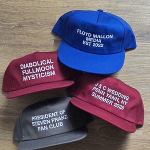 May include: Four baseball caps in various colors: blue, burgundy, and gray. Each cap features white embroidered text. The blue cap reads "FLOYD MALLON MEDIA EST 2022." The burgundy caps say "DIABOLICAL FULLMOON MYSTICISM" and "J & C WEDDING PENN YANN, NY SUMMER 2026." The gray cap reads "PRESIDENT OF STEVEN FRANZ FAN CLUB."