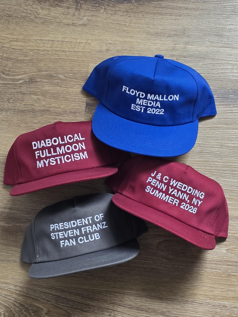 May include: Four baseball caps in various colors: blue, burgundy, and gray. Each cap features white embroidered text. The blue cap reads "FLOYD MALLON MEDIA EST 2022." The burgundy caps say "DIABOLICAL FULLMOON MYSTICISM" and "J & C WEDDING PENN YANN, NY SUMMER 2026." The gray cap reads "PRESIDENT OF STEVEN FRANZ FAN CLUB."
