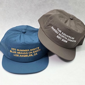 Puede incluir: Una gorra de béisbol azul con texto amarillo que dice HOT SUMMER NIGHTS BLUE HEAVAN ON EARTH LOS ANGELES, CA y una gorra gris con texto blanco que dice THE AQUARIUM FARGO NORTH DAKOTA EST 2006. Ambas gorras tienen corona y visera.