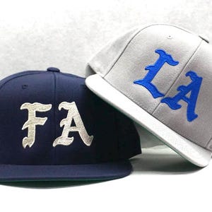 Puede incluir: Dos gorras de béisbol, una azul marino con letras blancas "FA" y otra gris con letras azules "LA".