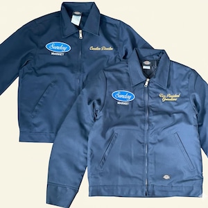 May include: Two navy blue work jackets with a zip-up front, collared neckline, and front pockets. One jacket has "Sunday Market" embroidered on the chest and "Creative Direction" on the right chest. The other jacket has "Sunday Market" and "Vice President Operations" embroidered.
