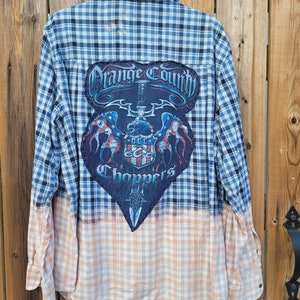 May include: Blue and white plaid button-down shirt with a large patch on the back that says "Orange County Choppers". The bottom half of the shirt is a faded peach and white plaid. The shirt is hanging on a wooden background.