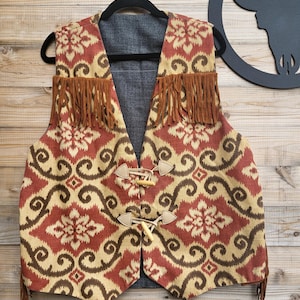 May include: A sleeveless waistcoat featuring a red and tan damask pattern with brown fringe detailing across the chest. The garment includes two light-coloured toggle closures on the front and is displayed on a black hanger against a wooden background.