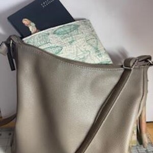 May include: A taupe-colored shoulder bag with a long strap, holding a black book and a patterned pouch. The pouch features a light green and white hot air balloon design. The book has the text "Collins Classic" on the cover.