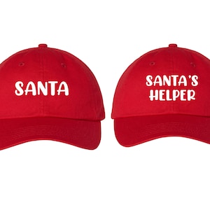 May include: Two red baseball caps with white text. One cap says "SANTA" and the other says "SANTA'S HELPER".