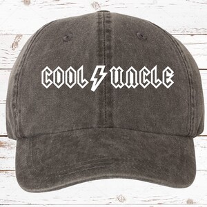 May include: A dark gray denim baseball cap with a white embroidered design that says "COOL UNCLE" with a lightning bolt in between the words.
