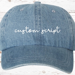 May include: A blue denim baseball cap with white embroidered text that reads "custom script".
