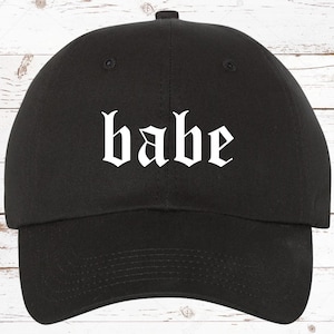 May include: Black baseball cap with the word "babe" in white gothic font across the front. The cap has a curved brim and a fabric strap closure on the back. The hat is made of a durable, solid-colored fabric.
