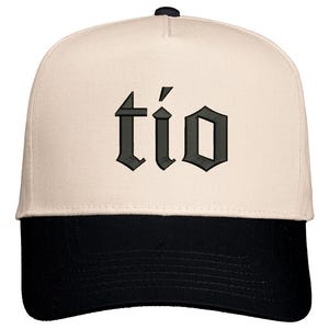 May include: A baseball cap with a cream-colored crown and a black brim. The word "tío" is embroidered in a dark gray gothic font across the front of the cap. The cap has a curved brim and a button on top.