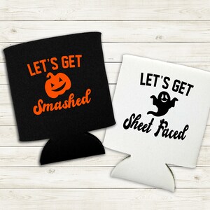 May include: Two black and white Halloween-themed can coolers. The black cooler has an orange pumpkin with a face and the text "Let's Get Smashed". The white cooler has a black ghost with a face and the text "Let's Get Sheet Faced".