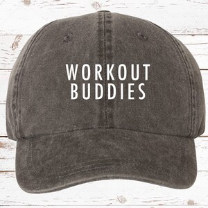 以下が含まれることがあります： 白い文字で「WORKOUT BUDDIES」と書かれたグレーの野球帽。つばがカーブしており、カジュアルな服装に適した、クラシックでアンストラクチャードなデザインです。