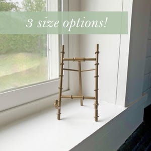 May include: A gold metal easel stand with three legs and a round top. The stand is on a white surface. 3 size options!