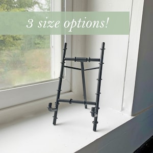 May include: Black metal easel stand with three size options. The easel is on a white window sill.