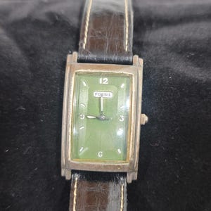 May include: A rectangular Fossil wristwatch featuring a green dial with silver-tone hour markers, the number 12, and a central Fossil logo. The watch has a silver-tone metal case and a dark brown textured leather strap with visible stitching.