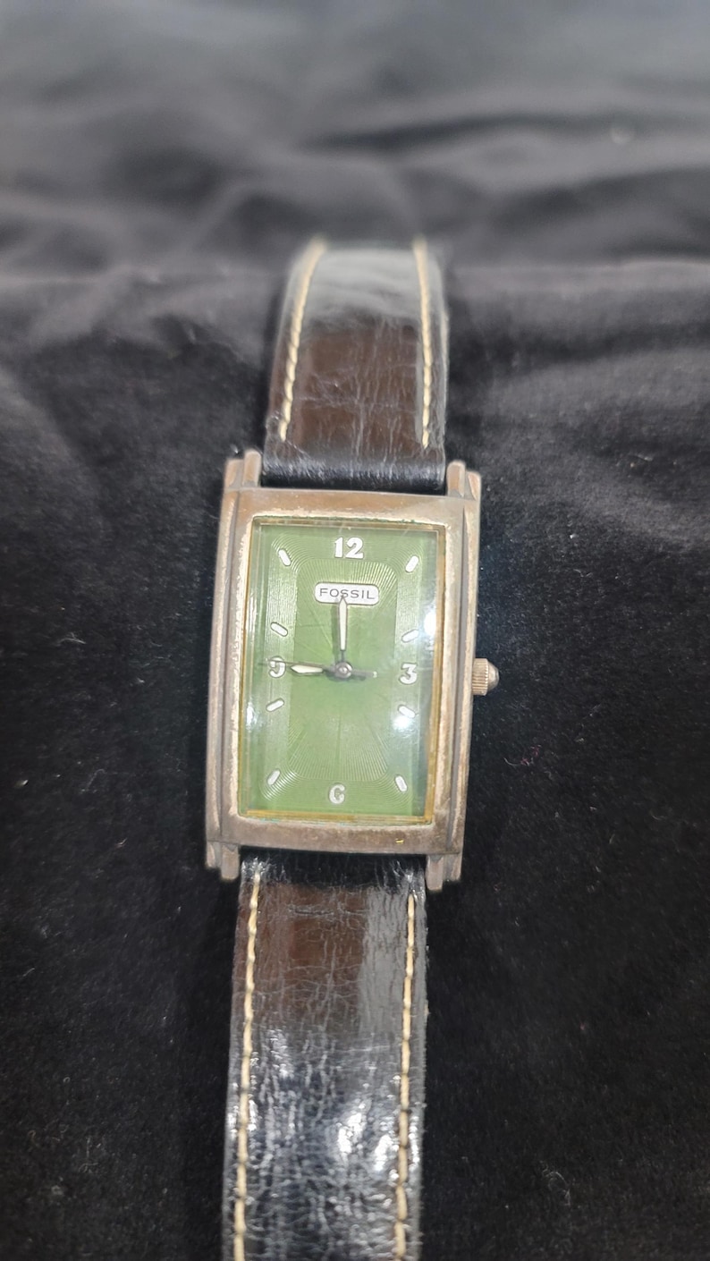 May include: A rectangular Fossil wristwatch featuring a green dial with silver-tone hour markers, the number 12, and a central Fossil logo. The watch has a silver-tone metal case and a dark brown textured leather strap with visible stitching.