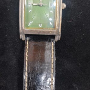 May include: A rectangular Fossil wristwatch featuring a green dial with silver-tone hands and markers. The watch has a metallic case with a weathered finish and a black leather strap with visible white stitching. The dial displays the number 12.