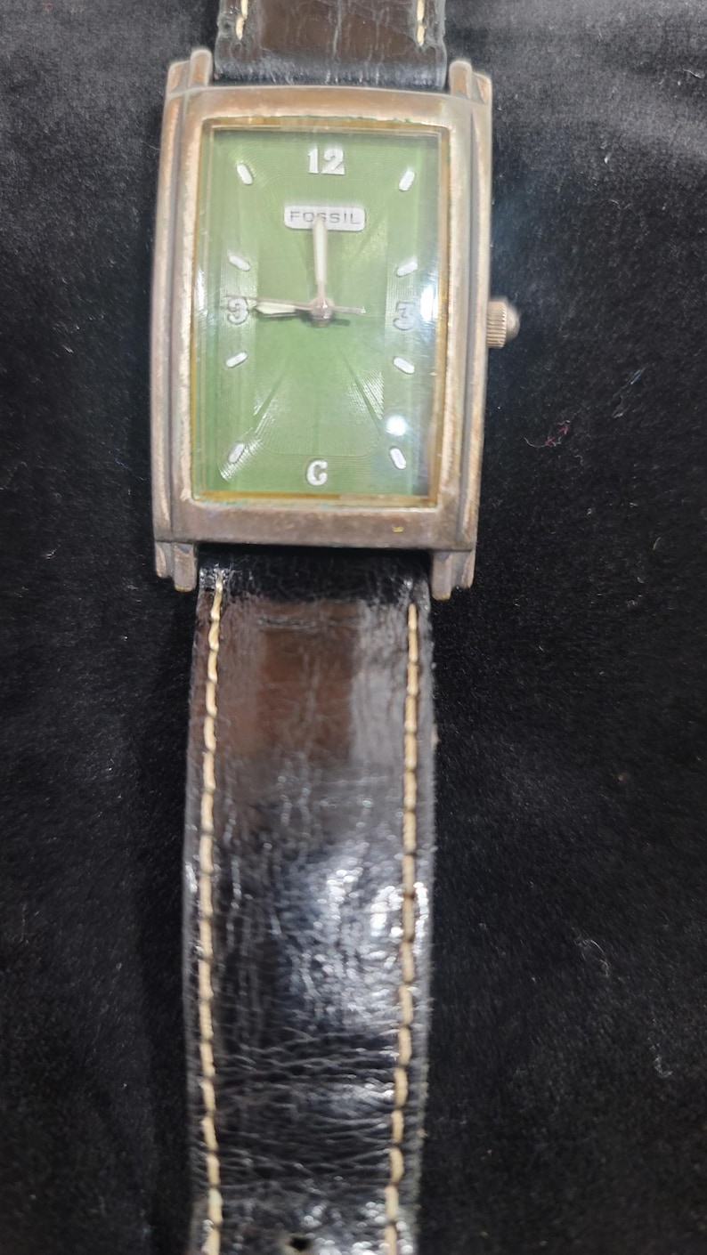 May include: A rectangular Fossil wristwatch featuring a green dial with silver-tone hands and markers. The watch has a metallic case with a weathered finish and a black leather strap with visible white stitching. The dial displays the number 12.