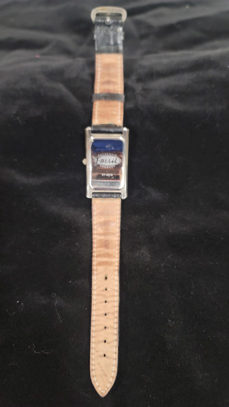 May include: A rectangular Fossil wristwatch featuring a silver-tone metal case and a black leather strap. The back of the watch case is metallic and displays the Fossil logo with the text Authentic Product and the model number VT-2578 clearly visible.