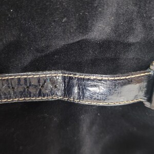 May include: A rectangular wristwatch featuring a dark green face and a black leather strap with visible light-colored stitching. The watch case is metallic, and the strap shows a textured, worn finish with multiple adjustment holes along the band.