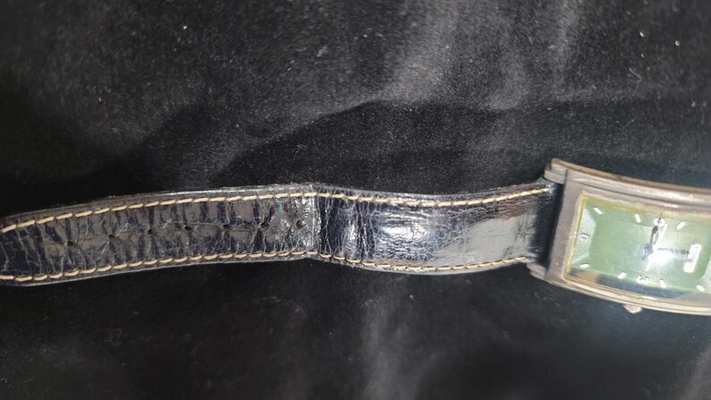 May include: A rectangular wristwatch featuring a dark green face and a black leather strap with visible light-colored stitching. The watch case is metallic, and the strap shows a textured, worn finish with multiple adjustment holes along the band.