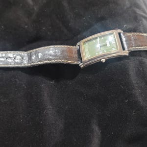 May include: A rectangular wristwatch featuring a dark green dial with silver-tone hands and hour markers. The watch has a polished silver-tone metal case and a dark brown textured leather strap with visible stitching along the edges.