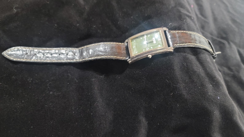 May include: A rectangular wristwatch featuring a dark green dial with silver-tone hands and hour markers. The watch has a polished silver-tone metal case and a dark brown textured leather strap with visible stitching along the edges.