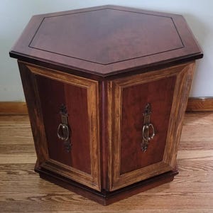 May include: A hexagonal wooden end table featuring a dark brown burl wood top and side panels with lighter wood trim. The piece includes two vertical cabinet doors, each adorned with a decorative antique brass drop pull handle centered on the panel.