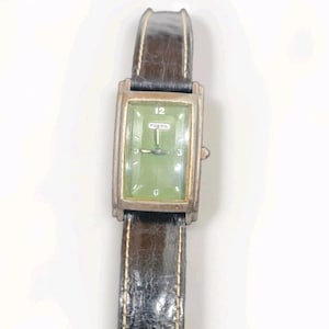 May include: A rectangular Fossil wristwatch featuring a green dial with silver-tone hands and markers, including the number 12 at the top. The watch has a metallic silver-tone case and a dark brown textured leather strap with visible light stitching.