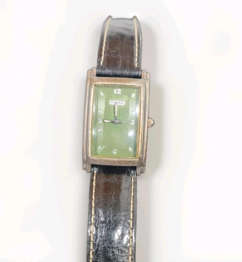 May include: A rectangular Fossil wristwatch featuring a green dial with silver-tone hands and markers, including the number 12 at the top. The watch has a metallic silver-tone case and a dark brown textured leather strap with visible light stitching.