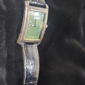 May include: A rectangular Fossil wristwatch featuring a green dial with silver-tone hands and markers, including the number 6. The watch has a silver-tone metal case and a black textured leather strap with visible stitching along the edges.