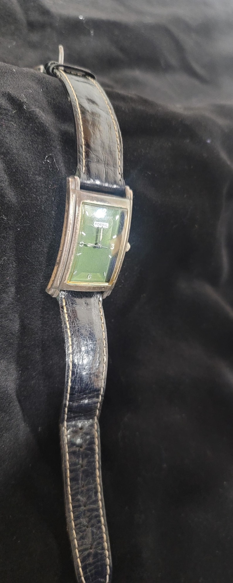 May include: A rectangular Fossil wristwatch featuring a green dial with silver-tone hands and markers, including the number 6. The watch has a silver-tone metal case and a black textured leather strap with visible stitching along the edges.