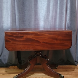 May include: A mahogany drop-leaf table with a rounded rectangular top and a pedestal base. The table has a rich, reddish-brown finish and stands on four curved legs with brass-colored claw feet. The table is set on a hardwood floor.
