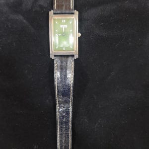 May include: A rectangular wristwatch with a green face, silver-tone metal casing, and a black leather strap with white stitching. The watch face features the number 12 at the top, the Fossil logo in the center, and a G at the bottom position.